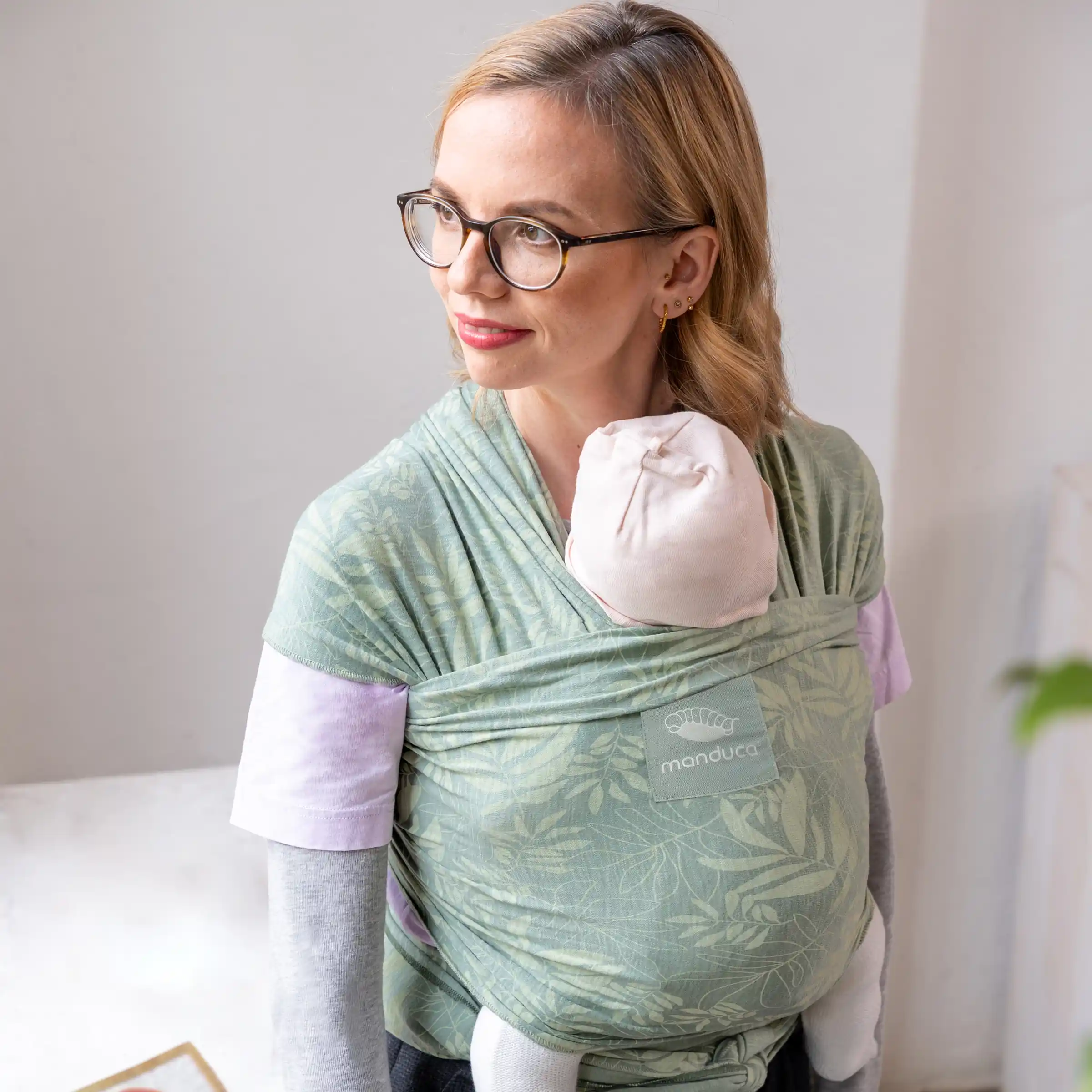 Frau mit Brille trägt ein Baby in einem grünen manduca Tragetuch und blickt zur Seite.